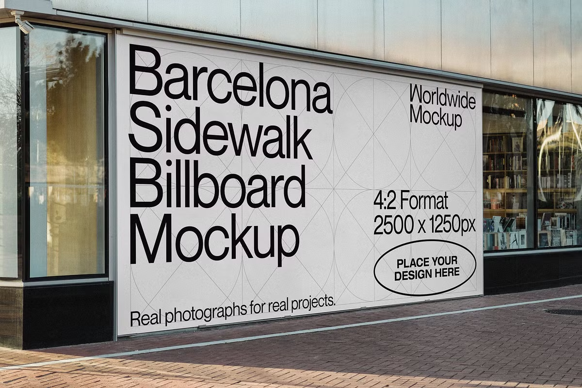 Mockup de um outdoor (billboard) instalado na fachada de um edifício, posicionado em uma calçada ao lado de uma vitrine de vidro. O painel apresenta textos técnicos em preto sobre um fundo cinza claro com grid de linhas geométricas: 'Barcelona Sidewalk Billboard Mockup', 'Worldwide Mockup', '4:2 Format' e '2500 x 1250px'. Um selo oval no canto inferior direito contém a instrução 'PLACE YOUR DESIGN HERE', exemplificando a aplicação de uma peça publicitária em contexto urbano.
