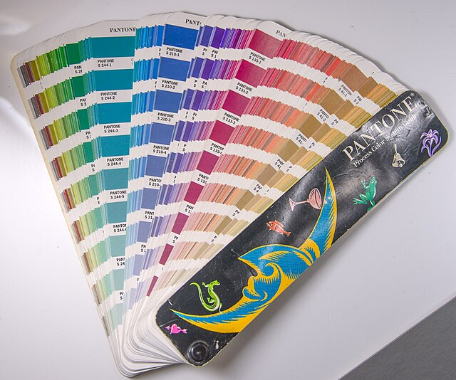 Exemplificação do sistema 'Pantone' através de um leque de cores aberto sobre uma superfície branca. A imagem mostra diversas lâminas com amostras de cores organizadas em degradês de verde, azul, roxo e rosa, cada uma com seu código de identificação. O texto no topo define o termo como um sistema internacional de padronização de cores para impressão por meio de tintas pré-misturadas (escala spot).