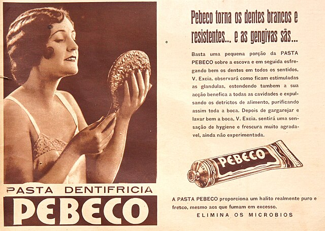 Exemplificação do termo 'Publicidade' através de um anúncio antigo da 'Pasta Dentifricia PEBECO'. A imagem, em tons de sépia, mostra uma mulher admirando o sorriso em um espelho de mão à esquerda, acompanhada de textos promocionais e a ilustração de um tubo de pasta de dentes à direita. No topo, o texto define publicidade como ações de comunicação que visam promover produtos ou serviços para obter aceitação do público.